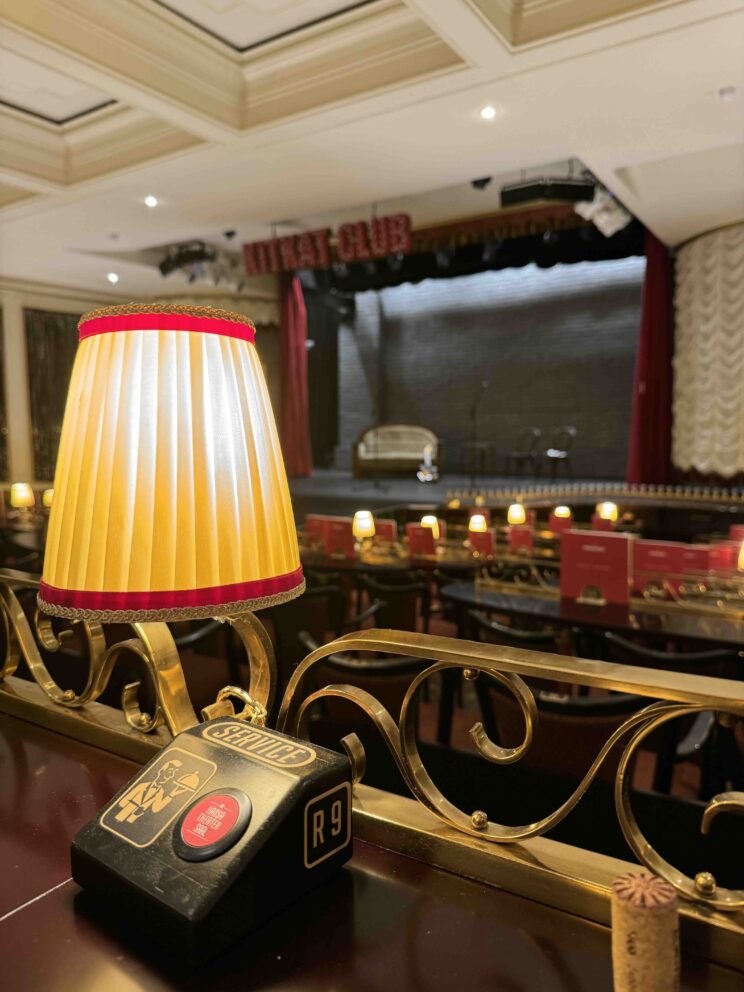 Close-Up einer Tischlampe auf einem Tisch mit Sitzplatz im Theatersaal, im Hintergrund weitere Tische und Sitze sowie die Bühne. Der Saal ist leer.