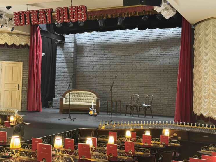 Blick in den leeren Theatersaal des Hansa-Theaters. Sitze mit kleinen Tischen, Tischlampen, auf der Bühne ein Sofa und zwei Stühle.